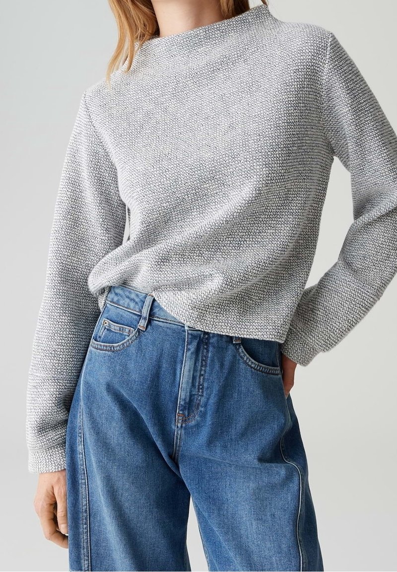 Femme portant un pull gris clair texturé, rentré dans un jean denim bleu ample à taille haute, sur un fond uni.