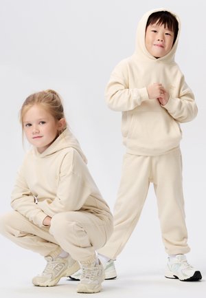 Beige hooded sweaters en joggingbroeken gemaakt van zachte stof; de jongen staat en het meisje gaat in knieligging, beide in bijpassende outfits met sneakers.