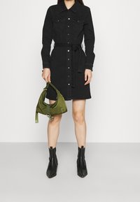 Vestido camisón de denim negro con cierre de botones y bolsillos frontales, ajustado en la cintura con un cinturón a juego. Combinado con un bolso verde y botas de tobillo negras.