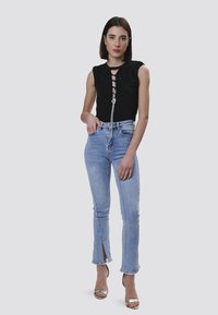 Top negro ajustado con detalle de ojal en la parte delantera, combinado con jeans skinny de color azul claro con dobladillos con abertura en la parte delantera y sandalias de tacón plateadas.