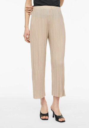 Kvinde iført beige plisserede viddebukser og sorte åbne høje hæle mule, stående mod hvid baggrund.