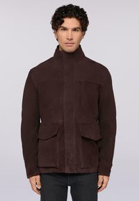 Giacca in suede marrone con colletto alto, zip frontale e due grandi tasche a pattina. Texture liscia con finitura opaca e dettagli minimi.