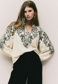 Bluză albă cu broderie florală neagră, decolteu în V și mâneci lungi și vaporoase, asortată cu pantaloni negri cu talie înaltă.