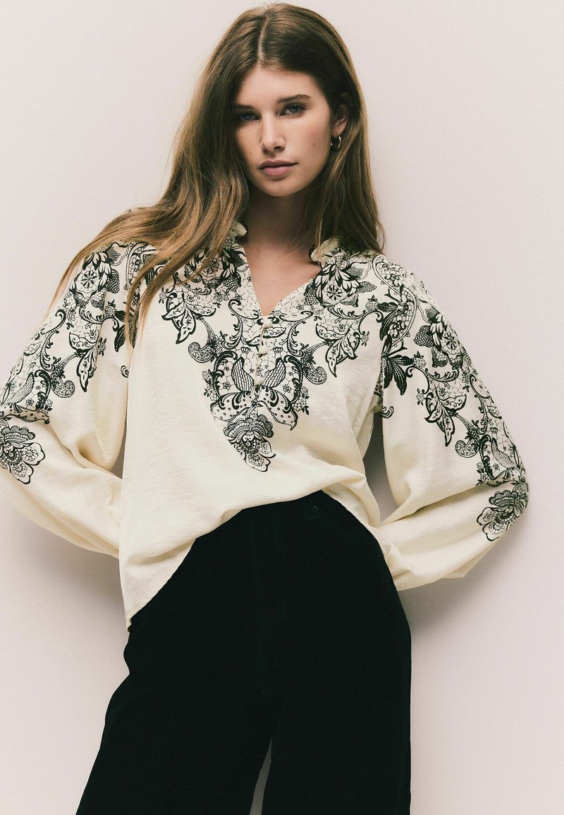 Bluză albă cu broderie florală neagră, decolteu în V și mâneci lungi și vaporoase, asortată cu pantaloni negri cu talie înaltă.