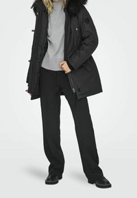 Parka noire matelassée avec une capuche doublée de fourrure, dotée de boutons-pression, de poches latérales et d'une taille cintrée, portée avec un pull gris et un pantalon noir.