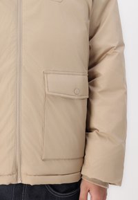 Beige pufferjakke med glidelås, en knappet klafflomme og quiltet tekstur på stoffet, med ribbestrikkede mansjetter på ermene.