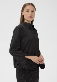 Crna bluzica s okovratnikom, sa perlicama na ovratniku i rukavima, izrađena od glatkog materijala i labavog kroja.