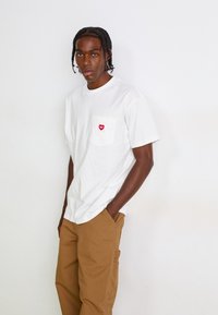 Young man with braided hair wearing a white T-shirt with a red heart logo and brown pants, standing against a plain white background.