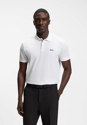 Mand iført en hvid kortærmet polo med "BOSS"-logo og sorte bukser, stående med den ene hånd i lommen mod ensfarvet baggrund.