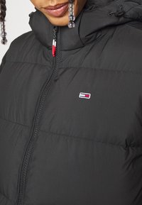 Svart puffjacka med hög krage, framdragkedja och liten logotyp med röd, vit och blå accent på bröstet. Slät tekstur.