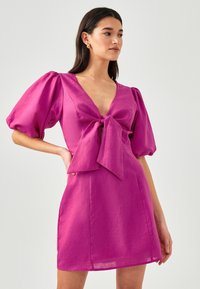 Fuksia lyhyttä mekkoa, jossa on syvä V-pääntie, puffihihat ja edessä nauhasolmu. Valmistettu kiiltävästä kankaasta, jossa on sileä tekstuuri ja tiukka muoto.