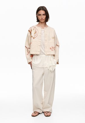 Mujer con chaqueta abotonada color crema bordada sobre una camiseta blanca, pantalones color crema de pierna ancha y sandalias marrones, de pie contra un fondo blanco.