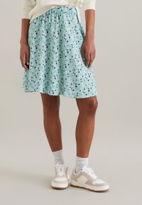 Jupe bleu clair avec un motif floral en vert, noir et blanc ; taille élastique ; longueur genoux avec un design évasé. Portée avec des baskets blanches.