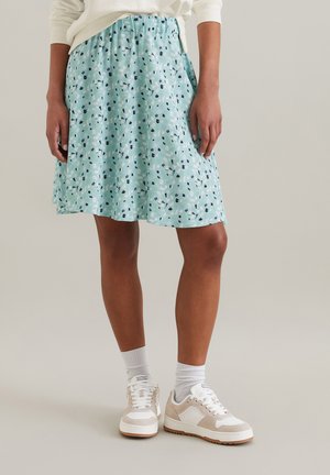 Hellblauer Rock mit floralem Muster in Grün, Schwarz und Weiß; elastischer Bund; knielang mit ausgestelltem Design. Kombiniert mit weißen Sneakers.