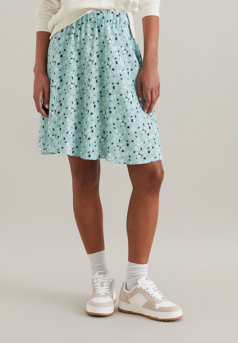 Jupe bleu clair avec un motif floral en vert, noir et blanc ; taille élastique ; longueur genoux avec un design évasé. Portée avec des baskets blanches.