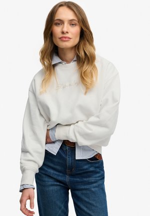 Frau mit langen, welligen Haaren trägt weißen Pullover über einem blau gestreiften Hemd, das in blaue Jeans mit braunem Gürtel gesteckt ist, vor einfachem Hintergrund.