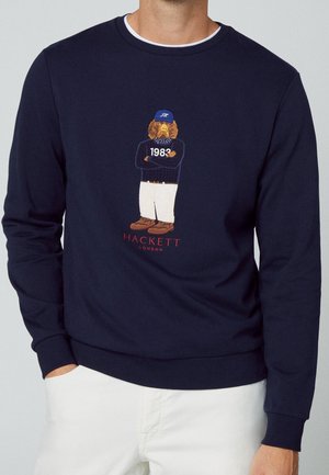 Granatowa bluza z haftowanym psem w niebieskiej czapce, sweter z "1983," białymi spodniami, brązowe buty i tekst "Hackett London" poniżej.
