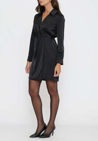 Abito avvolgente in satin nero con maniche lunghe, colletto e dettagli plissettati. Abbinato a scarpe con tacco alto nere e collant neri trasparenti.