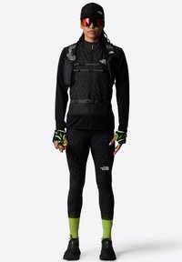 Svart zip-up jacka med ett texturerat mönster, figurnära svarta leggings, neongröna detaljer på strumpor och svarta träningsskor, komplett med en vätskeblåsa.