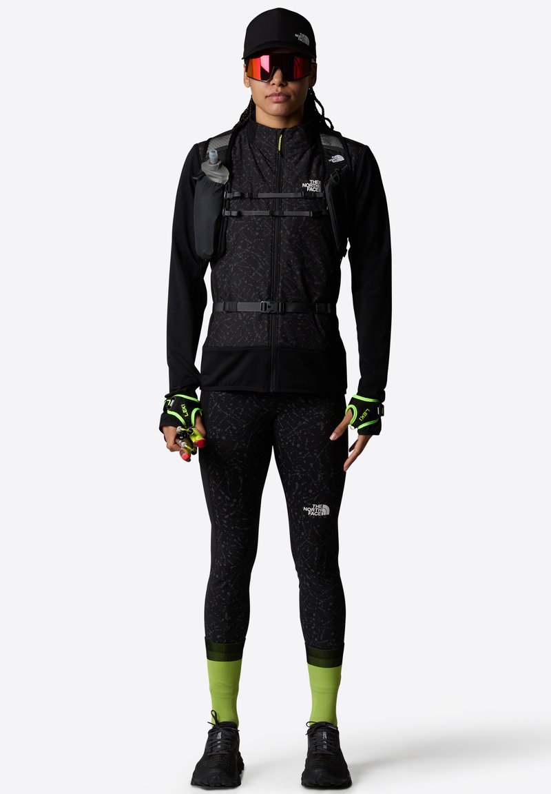 Svart zip-up jacka med ett texturerat mönster, figurnära svarta leggings, neongröna detaljer på strumpor och svarta träningsskor, komplett med en vätskeblåsa.