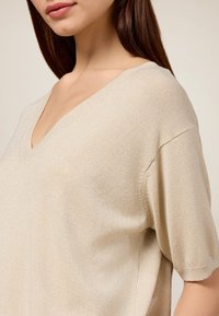 Beige Kurzarmoberteil mit V-Ausschnitt, aus einem strukturierten Strickmaterial mit dezentem Glitzereffekt. Nahtdetails an den Ärmeln und an der Seite.