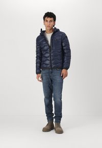 Giacca puffer navy con imbottitura texturizzata, chiusura con zip e cappuccio. Abbinata a jeans azzurri chiari e stivali beige con lacci.