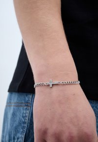 Stříbrný řetízkový náramek s malým akcentem kříže, do kterého jsou zasazeny čiré drahokamy. Řetěz má kovový vzhled a jednoduchý design.