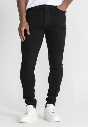 Person trägt schlanke schwarze Jeans und weiße Plateausneaker und steht mit einer Hand in der Tasche vor einfachem Hintergrund.
