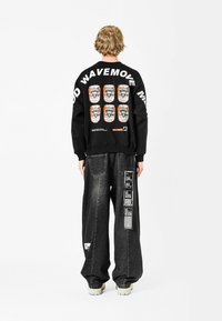 Sudadera negra con estampado gráfico en la parte trasera, que presenta seis latas y texto. Combinada con pantalones de denim oscuros y holgados, que incluyen parches laterales y etiquetas.