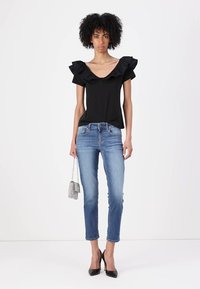 Top nero a maniche corte con spalline arricciate, abbinato a jeans aderenti azzurri e tacchi alti neri; tiene in mano una piccola borsa argentata.