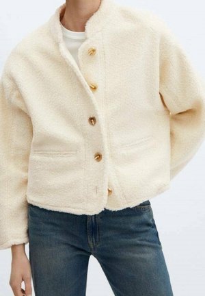 Fløjlsjakke i cremefarvet med knapper og bred krave båret over en hvid top, kombineret med højttaljede blå jeans.