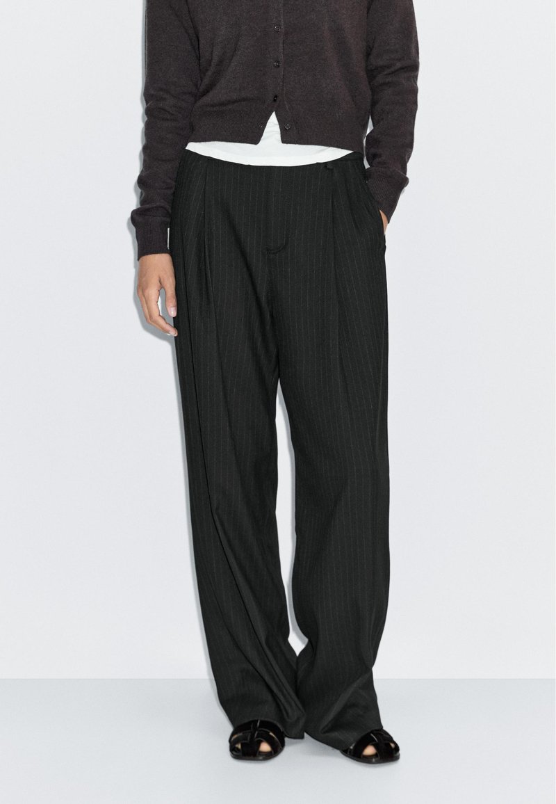 Pantaloni a gamba larga neri con righe sottili e vita alta, con pieghe frontali e tasche laterali. Abbinati a un cardigan scuro e sandali.