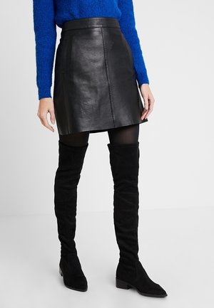 Frau trägt schwarzen Leder-Minirock, schwarze Strumpfhosen, schwarze Overknee-Stiefel und einen blauen Langarm-Pullover, steht vor weißem Hintergrund.