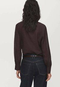 Chemise à manches longues bordeaux foncé en tissu texturé, avec des poignets boutonnés et une coupe décontractée, associée à un pantalon en denim bleu foncé et une ceinture marron.