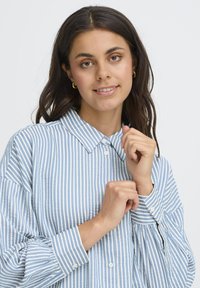 Young woman with dark hair wearing gold hoop earrings and a blue-and-white striped button-up shirt, adjusting her collar and cuffs.