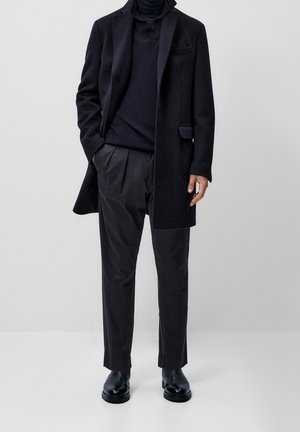 Manteau en laine navy sur un col roulé navy, associé à un pantalon plissé noir et des bottines noires. Texture lisse avec un design ajusté.