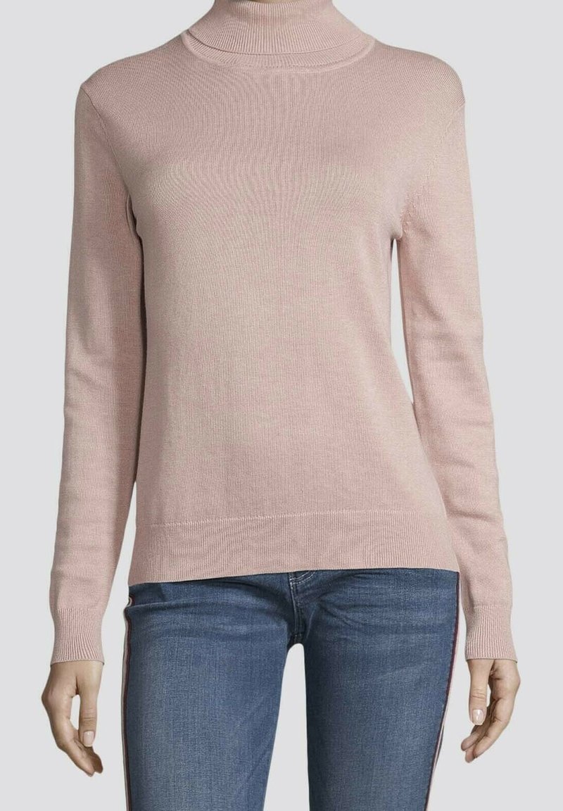 Pull à col roulé rose pâle à manches longues associé à un jean bleu avec des bandes rouges sur les côtés, porté par une personne sur un fond uni.
