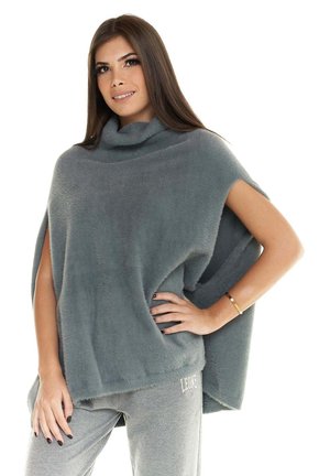 Une femme pose devant un fond blanc, vêtue d’un poncho gris duveteux et d’un pantalon assorti, une main sur la hanche.