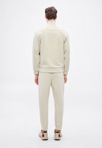 Beżowa bluza z wysokim kołnierzem i rękawami raglanowymi, zestawiona z dopasowanymi beżowymi dresami. Charakterystyczny detal kieszeni na spodniach.