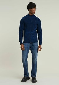 Maglione zip-up blu navy con design a maglia testurizzata, polsini a coste, indossato sopra jeans blu e scarpe nere, accessoriato con un berretto nero.