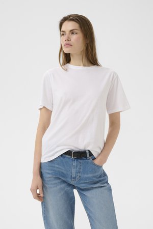 Nuori pitkähiuksinen nainen, joka pukeutunut yksinkertaiseen valkoiseen t-paitaan, joka on tungettu vaaleansinisiin farkkuihin mustan vyön kanssa, käsi taskussa, katsoo vasemmalle.