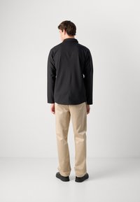 En mann står med ryggen til, iført en svart langermet skjorte, beige bukser og svarte sko mot en ensfarget hvit bakgrunn.