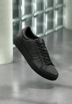 Black leather sneaker with a low profile, featuring black laces, textured accents, and a rubber sole. Logo embossed on outer side.