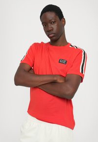 Red t-shirt with short sleeves, featuring black and white striped accents on shoulders and a small black logo patch on the chest.