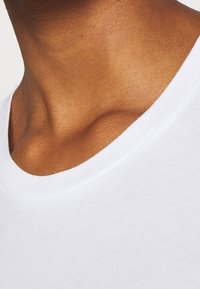 Biała bawełniana koszulka z okrągłym dekoltem, gładką fakturą oraz zbliżeniem na tkaninę i odcień skóry pod kołnierzem.