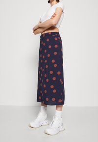 Laivansinisellä midi-hameella, jossa on suuria oransseja pilkkuja, on joustava vyötärö ja valuva kangas. Hame on yhdistetty valkoisiin paksupohjaisiin tennareihin.