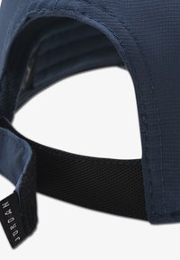 Καπέλο σε ναυτικό μπλε με υφή υφάσματος, διαθέτει μαύρο ρυθμιζόμενο λουράκι και μια μικρή ετικέτα με τη λέξη "JORDAN" σε λευκό χρώμα.