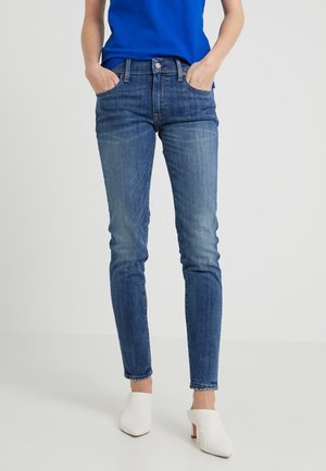 Persoon met slim-fit blauwe jeans, blauw overhemd en witte slip-on schoenen met hak, staand met handen in de zakken tegen een effen achtergrond.