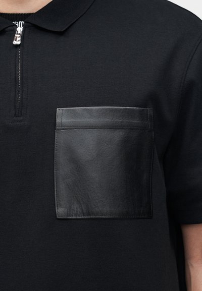 Μαύρο πόλο με φερμουάρ μπροστά και μεγάλη τετράγωνη μαύρη δερμάτινη τσέπη στο στήθος αριστερά.