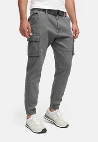 Homme portant un pantalon cargo gris avec des poches latérales, une ceinture noire et des baskets blanches, debout sur un fond blanc.